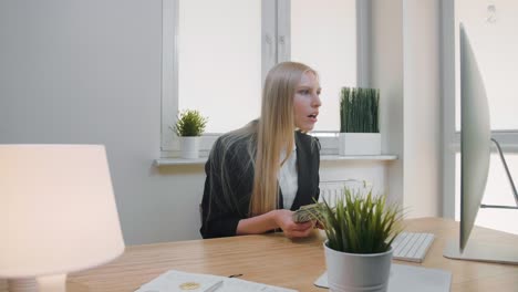 woman celebrating success in office. elegant blond female sitting at workplace holding in hands bundle of cash looking astonished at monitor, and throwing money in air and happily making yes gesture.