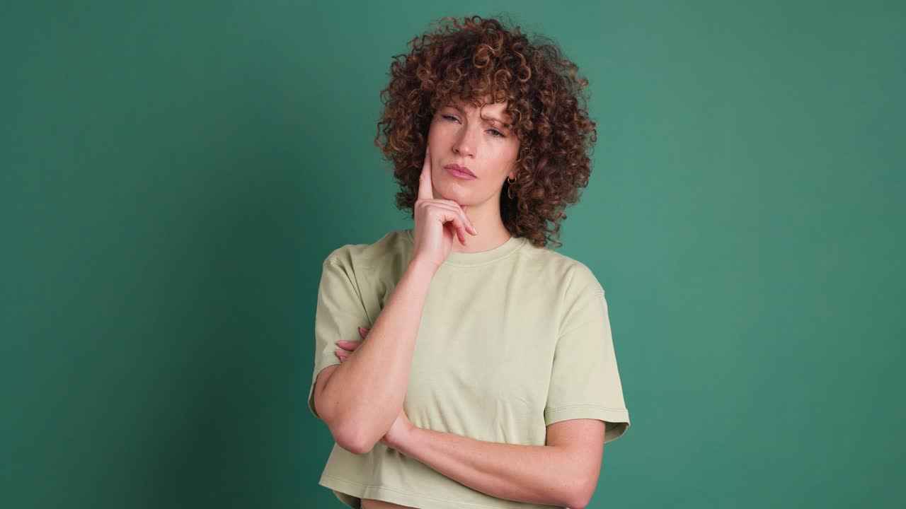 Thoughtful young woman touching face and smiling on green background