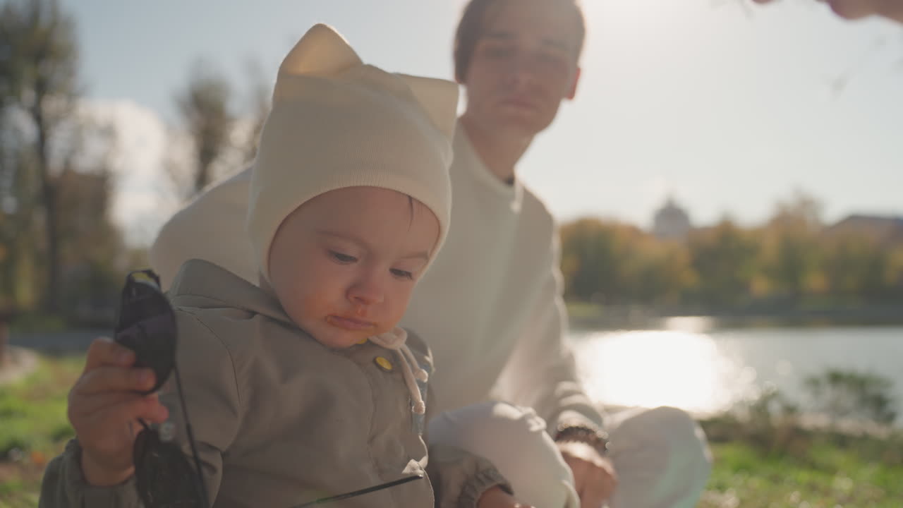 Solbeskinnet familiepicnic ved søen med en hvid baby iført strikhue, kærlige voksne læner sig tæt ind på hinanden på den grønne bred ved det reflekterende vand; en fredelig hyggescene, der indfanger en varm forbindelse og en ægteskabelig familieatmosfære.
