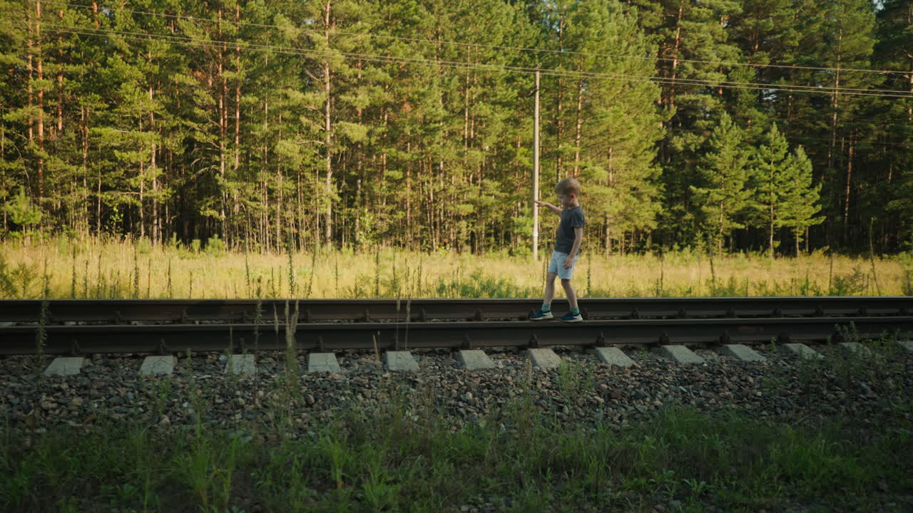 side view of young boy walking carefully on railway track beside grassy area with tall pine trees in background as he loses balance slightly while stepping down from rail beam