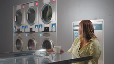 female entrepreneur carrying cup of tea and notebook walks to table inside laundromat, placing items as industrial washers with digital panels spin clothes in background, showcasing modern service