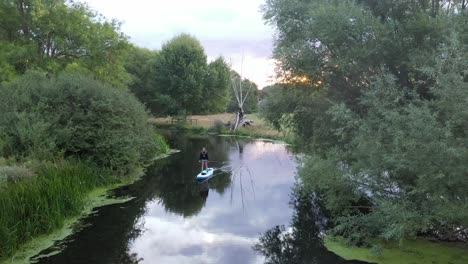 Woman-slowly-paddle-boarding-at-sunrise-along-the-river-stour,-Flatford,-Suffolk,-England