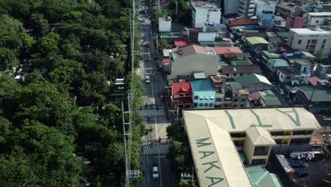 Drohnenaufnahme-Einer-Straße-Neben-Einem-Friedhof-Voller-Bäume-Und-Einer-Schule-Mit-Makati-Schrift-Auf-Dem-Dach