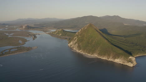 A-landscape-view-of-a-pyramid-shaped-mountain-located-at-a-river-estuary-flowing-into-the-sea,-with-green-vegetation-on-its-sides,-mountain-ridge-in-the-background,-on-the-sunset