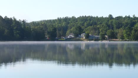 Morning-Handheld-View-of-Fog-Over-Lake---Small-Town-and-Forest-in-Background