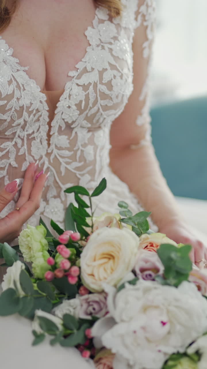 Stylish young bride sits on a couch and holds a beautiful tender flowers. Young model in white wedding dress is sitting in the light room. Vertical video