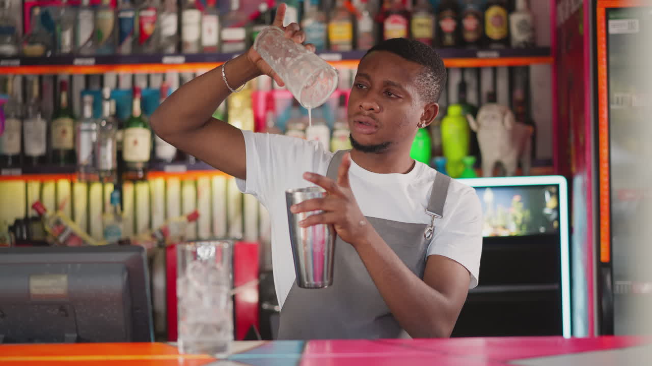 Bartender takes glass off shaker at counter. African American barman with ready refreshing drink in stainless cup against bar racks. Black servant work