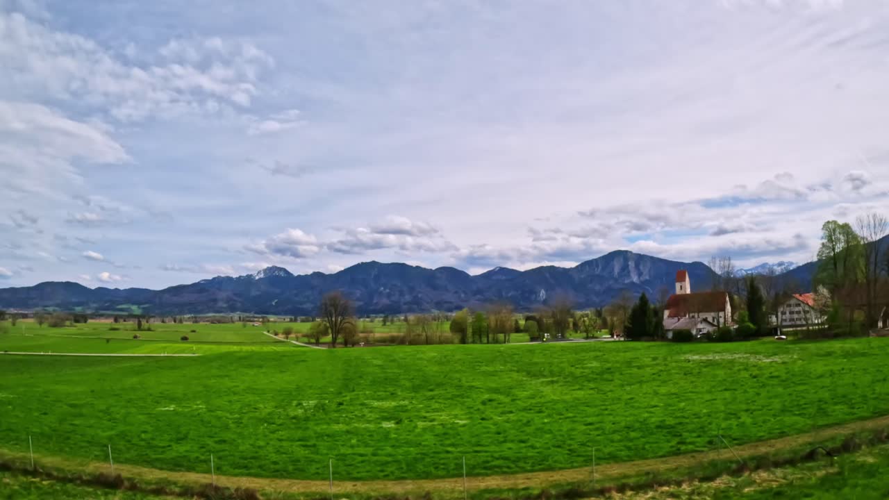 Extreme wide angle POV shot from a moving vehicle, open green meadows, rural lanes, and rolling foothills near the Austria-Germany border; scenic Alps in background