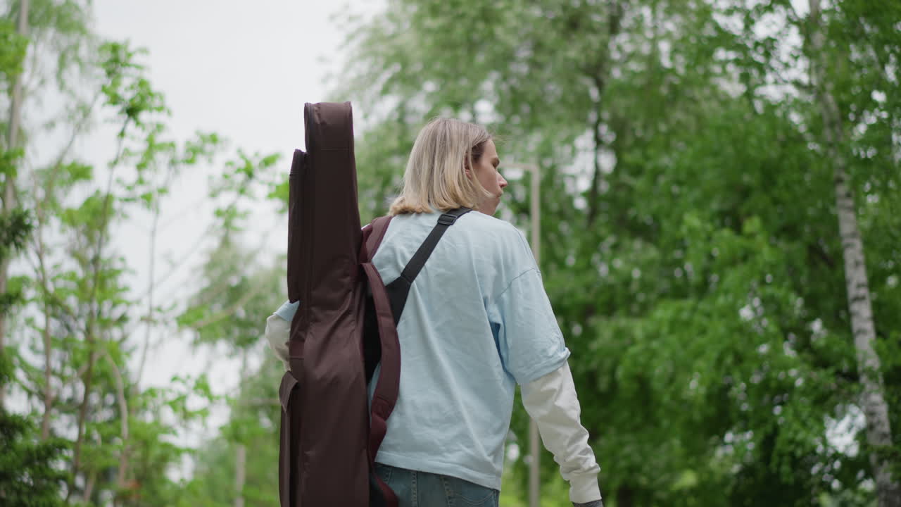 Un chitarrista percorre un sentiero nella foresta con una chitarra, atmosfera indie folk, cielo nuvoloso, chioma frondosa, camicia di jeans, lunghi capelli biondi, un musicista solista alla ricerca di idee per scrivere canzoni, si ferma ad ascoltare, riflessivo