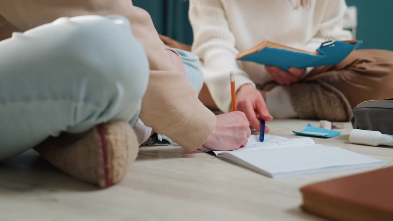 Primo piano di mani e gambe che scrivono, con tutor e studente caucasici seduti sul pavimento con quaderno e blocco appunti, segni di penna sulla carta, un tappeto accogliente e maglioni morbidi, luce naturale calda, in una tranquilla sessione di coaching grammaticale.