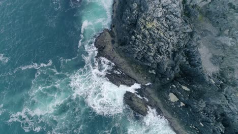 Antena-De-Arriba-Hacia-Abajo-De-Un-Escarpado-Acantilado-Rocoso-De-Color-Oscuro-Con-Poderosas-Olas-Rompiendo-Creando-Espuma-Del-Agua-Atlántica-Azul-Oscuro