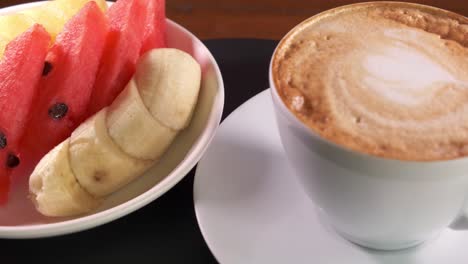 una taza de café caliente con espuma y un plato de fruta lateral girando en un plato