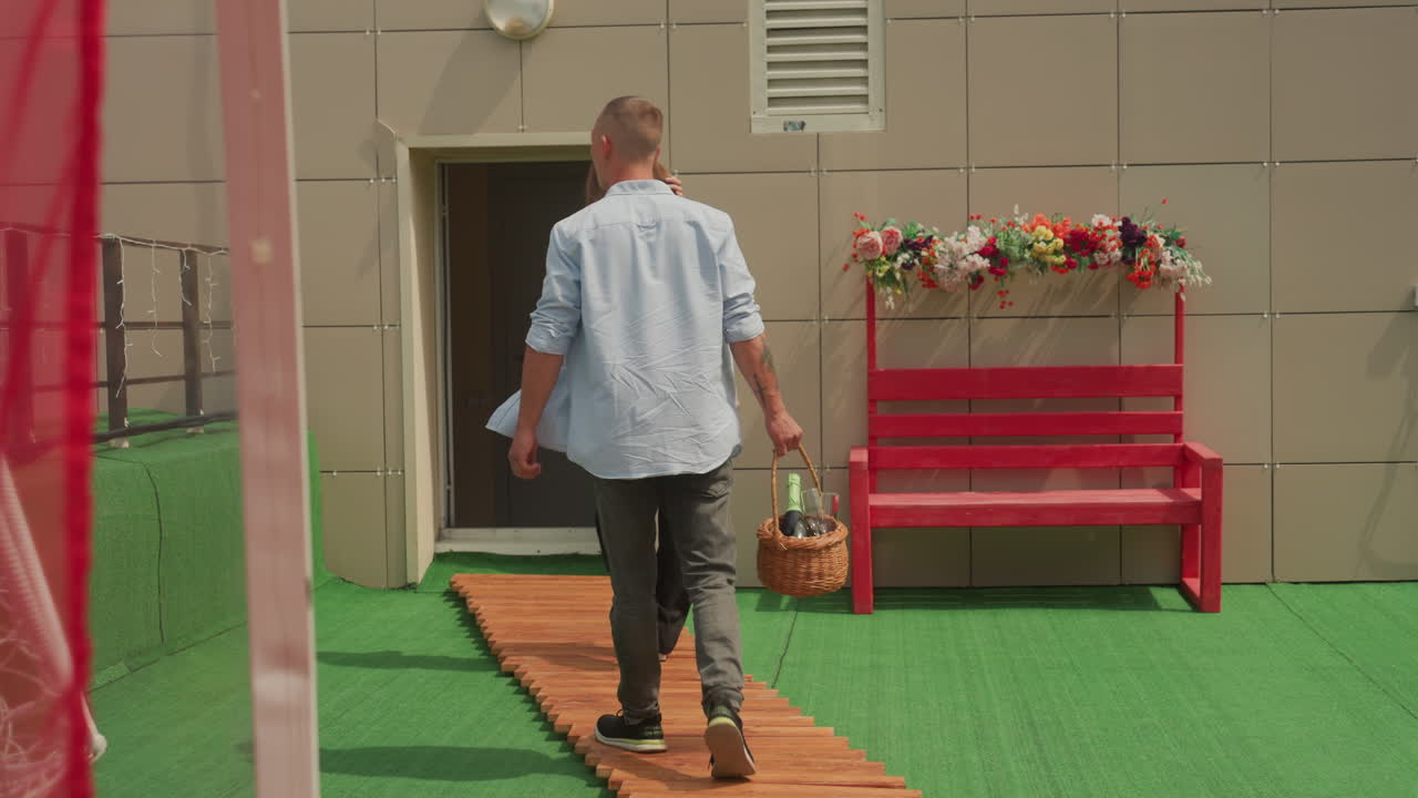 white couple walking on terrace bench man carrying woven basket with flowers while woman follows, sunny urban rooftop terrace with red bench, wooden path, casual summer clothing, visible tattoo