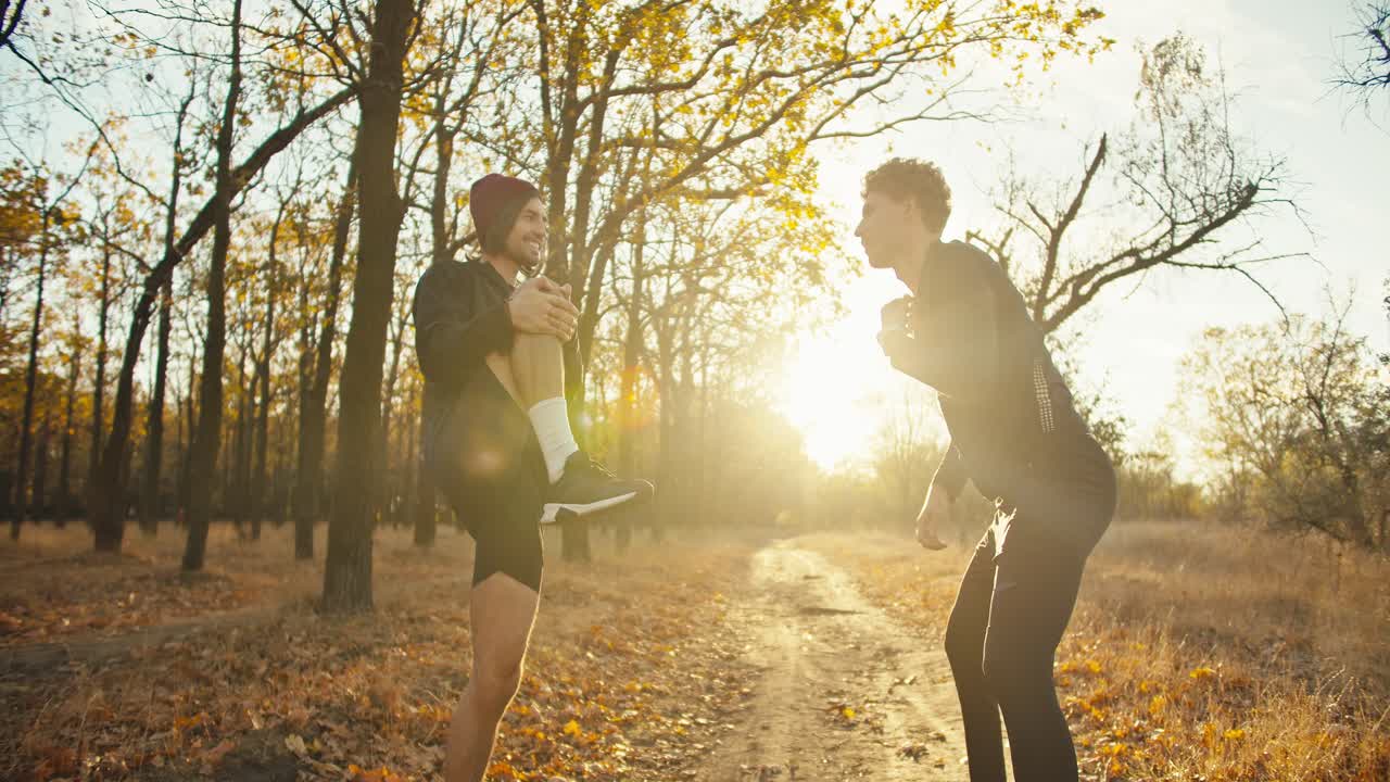 A man with curly hair in a black sports uniform together with his friend in black shorts and a red hat are doing sports and warming up before their jog in the autumn forest in the evening at sunset on an earthen path