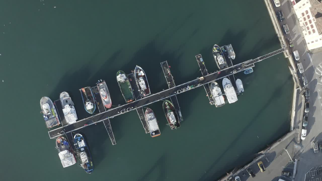 Fishing boats moored along pier in calm harbor water, Saint-Jean-de-Luz, France. Ideal for maritime, fishing industry concepts. Aerial top-down orbit descending