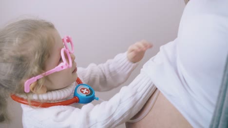 little girl in funny glasses plays doctor with pregnant mom