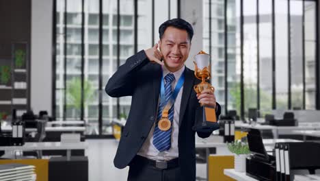 asian business man in a suit and tie with a gold medal and trophy showing call me gesture and smiling to camera as the first winner in office