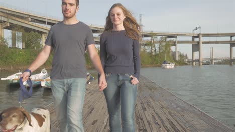 pannning shot of a young loving couple walking their adorable dog on a dock at sunset