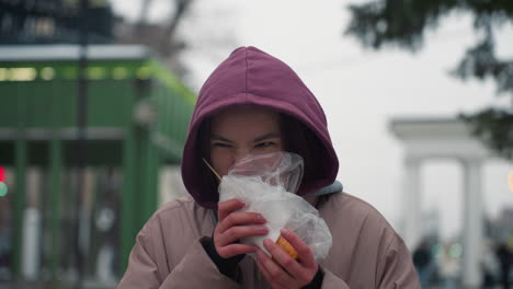 young woman in winter jacket eating pastry outdoors in urban park, holding wrapped food, cold weather, casual lifestyle, hood up, enjoying street food in winter season