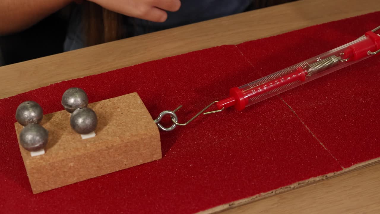 A hand uses a spring scale to measure force on a weighted block on sandpaper, illustrating friction concepts
