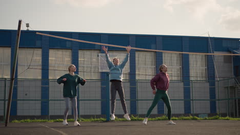 females in hoodies engage in volleyball training outdoors on court, background features basketball hoop, greenery, and building, women jumping with hands raised during practice session