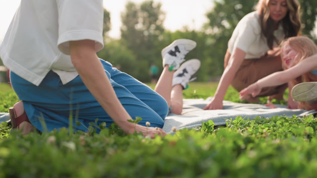 playful moms spreading blanket on green grass while children jump and laugh, summer park scene, sunlit family activity, joyful motion, close angle capturing connection and movement