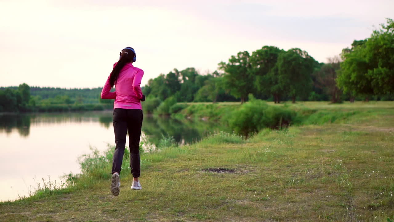 Ragazza mora con capelli lunghi, con le cuffie, corre lungo il fiume nel parco al mattino all'alba d'estate, con una giacca rosa e pantaloni neri