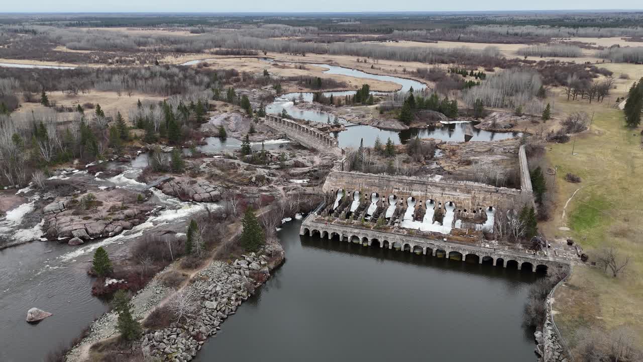 Drone luchtovervlucht van de historische Pinawa Dam in Manitoba’s Heritage Park tijdens de lente. Er ligt nog sneeuw op de grond, met omringend boreaal bos, een rivier en de oude ruïnes van de waterkrachtcentrale in zicht.