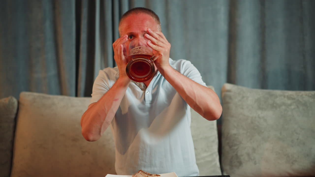 Middle aged man seated on sofa in indoor setting drinks from large transparent beer mug, pauses with intense gaze as he slowly lowers cup, soft background with curtains and pillows