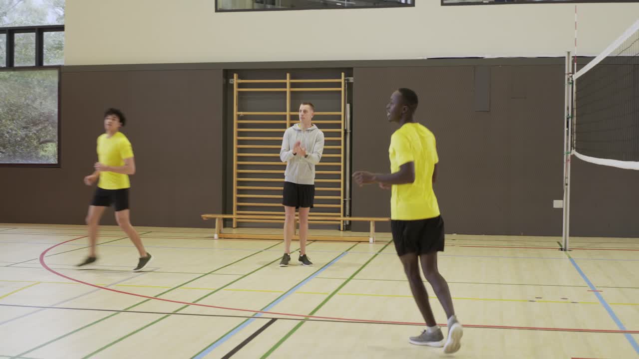 Time masculino diverso praticando vôlei perto da rede na academia, um jogador segurando uma bola colorida e depois arremessando-a.