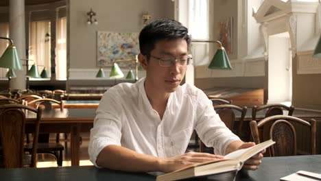 Young-asian-male-student-reading-book-and-smiling-at-camera-in-library