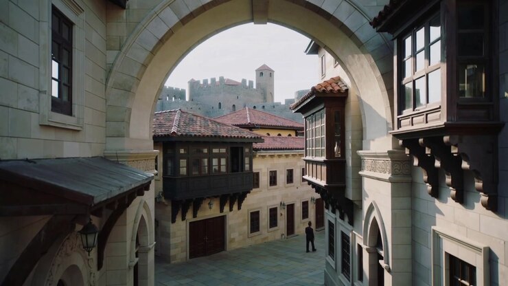 Inquadrando un arco di pietra, con la luce del sole che crea un bagliore, la telecamera rimane sotto, mentre un uomo vestito di scuro cammina verso destra.