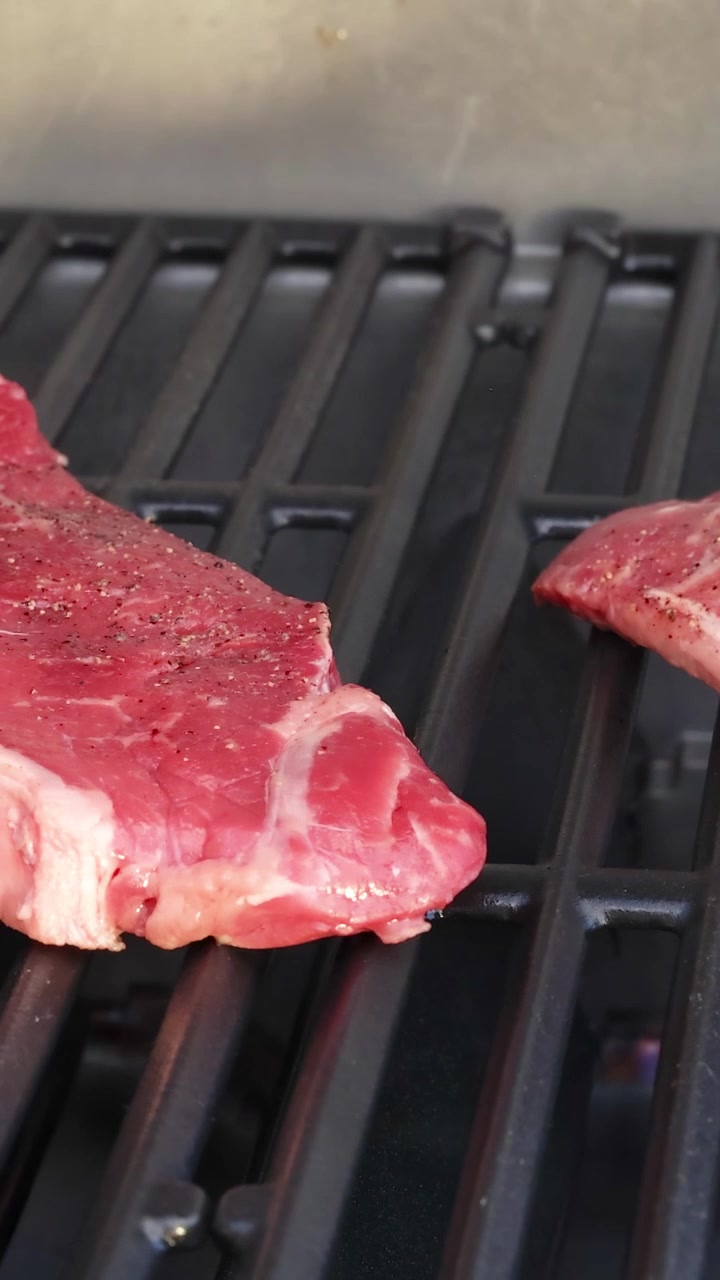 Raw beef steaks placed on a grill, beginning to cook over open flames. Close-up view with natural lighting