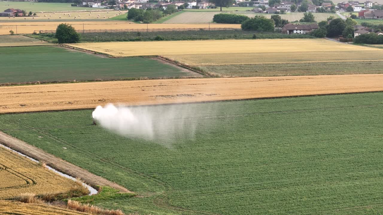 Aerial view revealing irrigation system spraying water across alfalfa and wheat fields in sunlit Po Valley landscape, PC, Italy