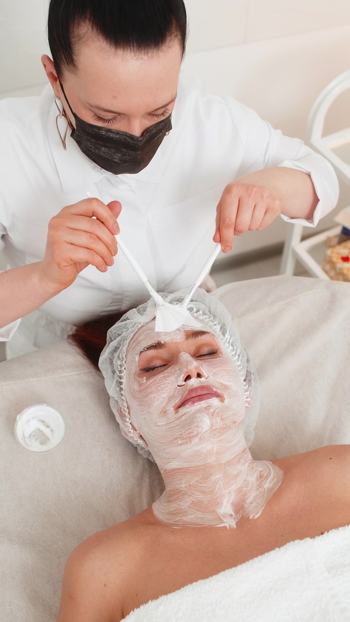 Estetista di terapia enzimatica che applica crema medica idratante al viso di una giovane donna adulta con i pennelli
