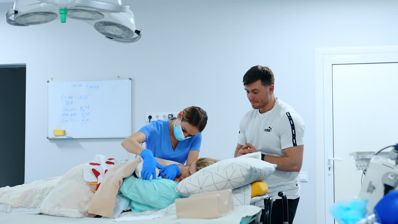 Female medic is helping mom to breastfeed her baby for the first time. Father is standing beside watching.