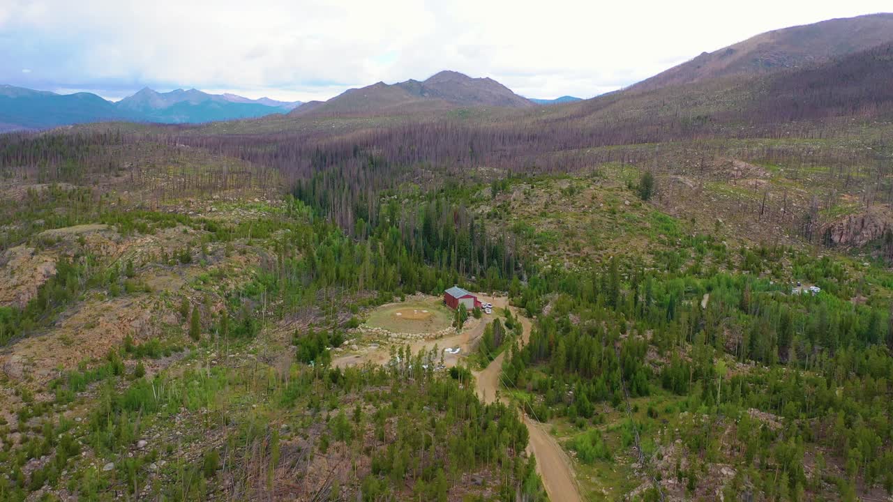 Vista aerea di una remota valle montana con foresta in ripresa, alberi morti, una strada sterrata tortuosa e una baita isolata circondata da aspro terreno montano sotto un cielo coperto in una vasta area selvaggia.