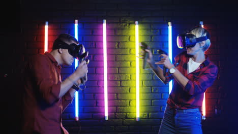 cheerful boyfriend and girlfriend in vr glasses playing virtual reality game and shooting with joysticks with neon colorful wall on the background