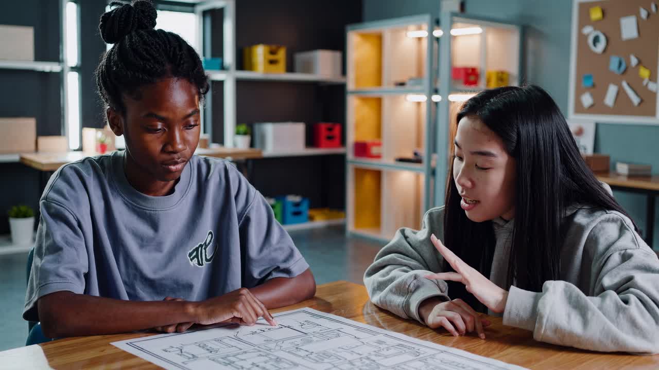 Two students study a blueprint at a table in a classroom. The video is shot from a side angle