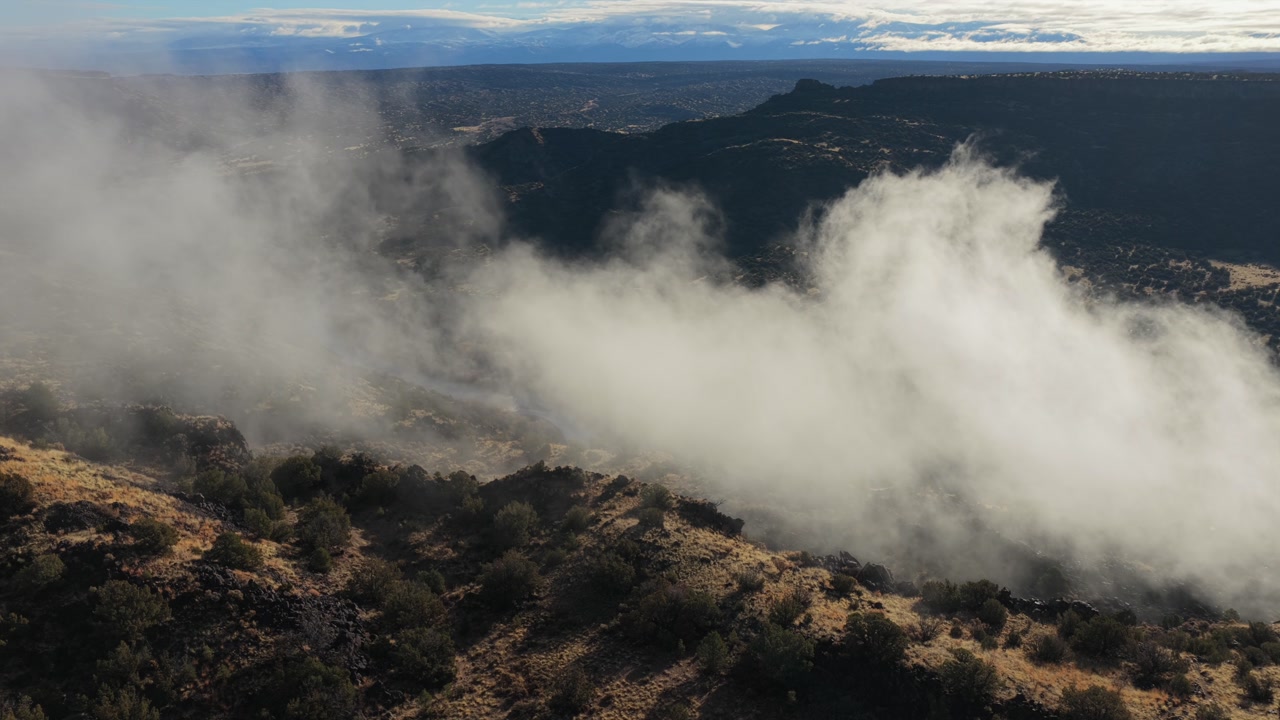 Drone shot shows thick white mist forming and rising from within a deep rugged wilderness canyon. The footage captures the atmospheric clouds moving over the rocky terrain and vegetation.