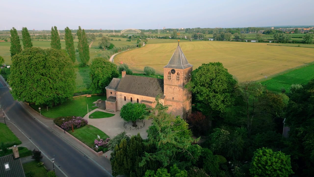 Luftaufnahme der historischen Oude Kerk in Oosterbeek, Niederlande, die ihre alte Architektur inmitten üppiger grüner Felder und Bäume unter einem heiteren blauen Himmel zeigt. Erfasst die ruhige Dorflandschaft.