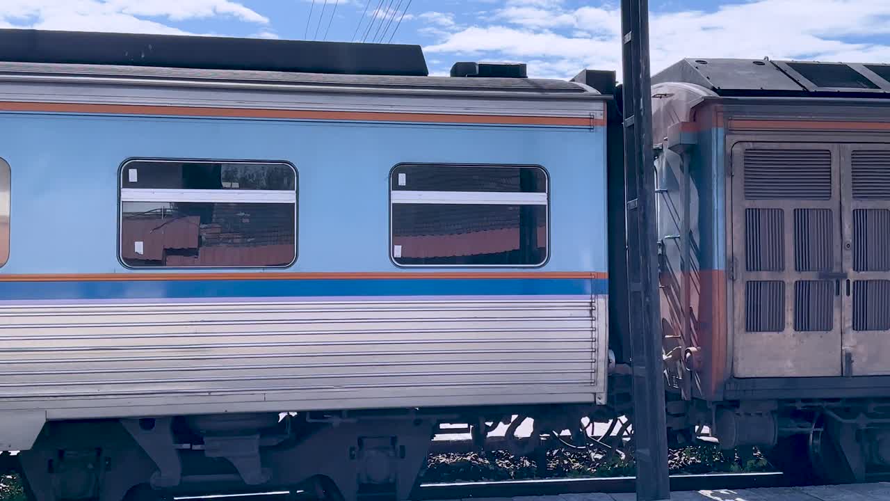 Side view of local passenger train slowly departing sunlit rural Thailand station, handheld tracking shot