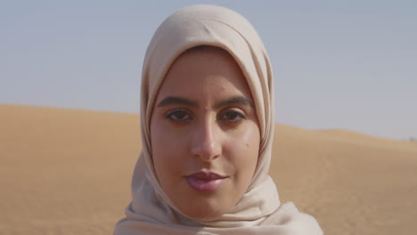 close up portrait of a beautiful muslim woman in hijab standing in a windy desert and looking at camera