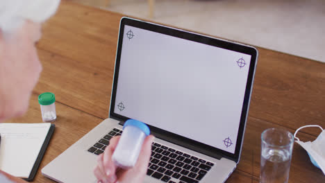 Senior-caucasian-woman-holding-medication-bottle-while-looking-at-laptop-screen-at-home