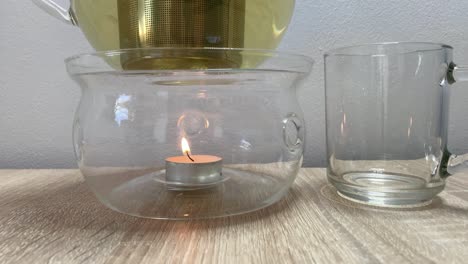 close-up on clear glass teapot warmer with small burning candle and tea cup