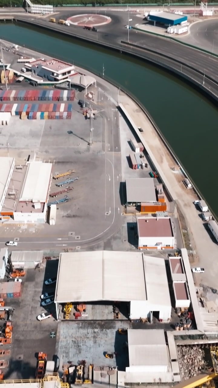 Terbang lintas udara dari atas Terminal Kontainer Manzanillo di Pelabuhan Manzanillo, Colima, Meksiko yang menghadap infrastruktur penanganan kargo, derek gantry, dan jalan layanan