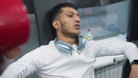 close up exhausted man leaning back on exercise machine after intense gym session wearing white workout top with wireless headphones around neck near window showing blurred outdoor view