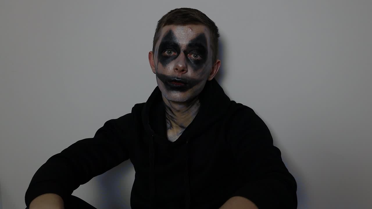 joven caucásico con maquillaje de halloween de payaso negro sentado y hablando con la cámara