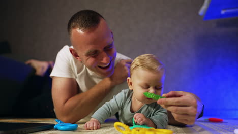 caucasian dad and his infant son lie on the floor. father gives a toy for teeth to his little baby.