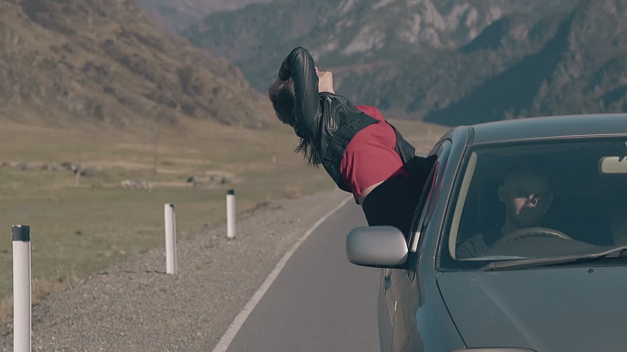 어두운 머리카락을 가진 예쁜 행복한 여자가 속도를 높이는 자동차 창에 앉아 있습니다.